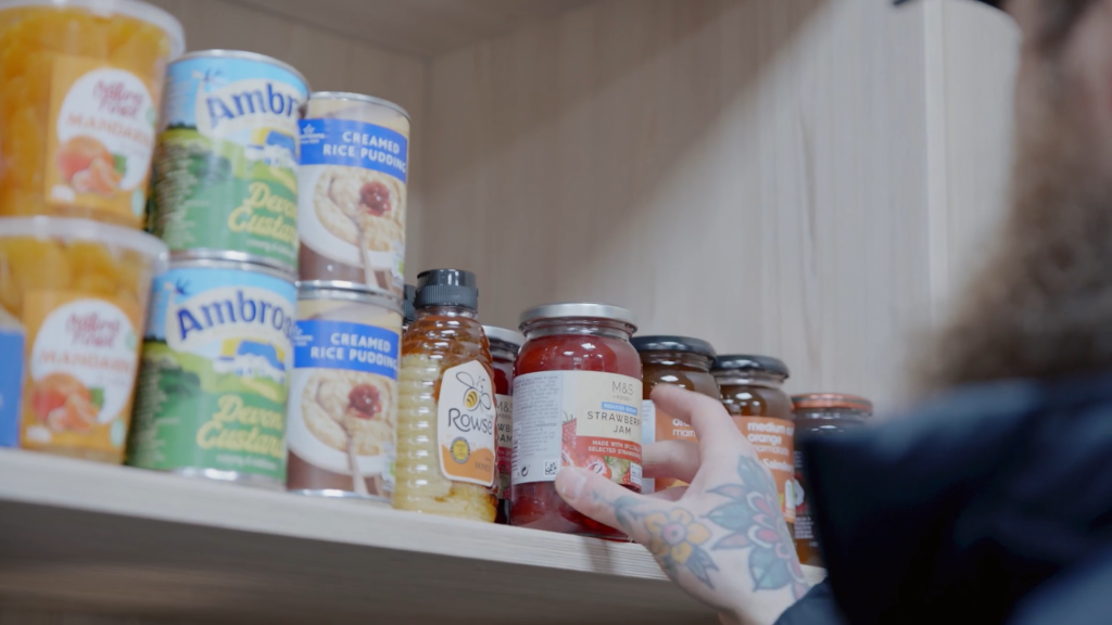 A hand reaches for a jar of jam on a shelf of tins and jars.