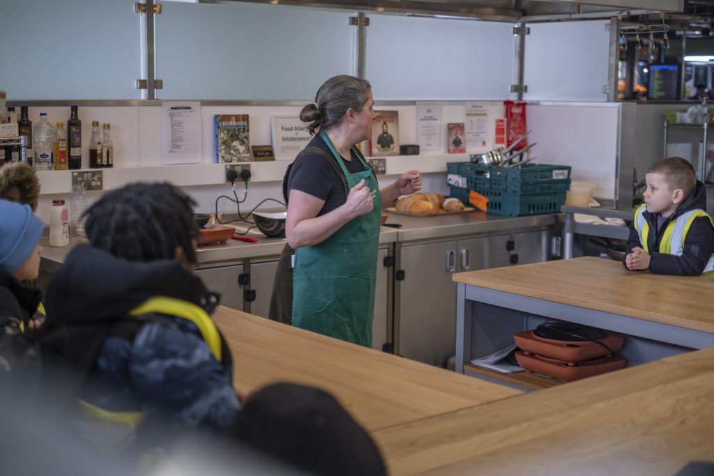 A chef explains cooking techniques to some young children sitting at tables.