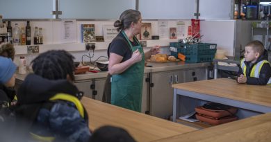 A chef explains cooking techniques to some young children sitting at tables.